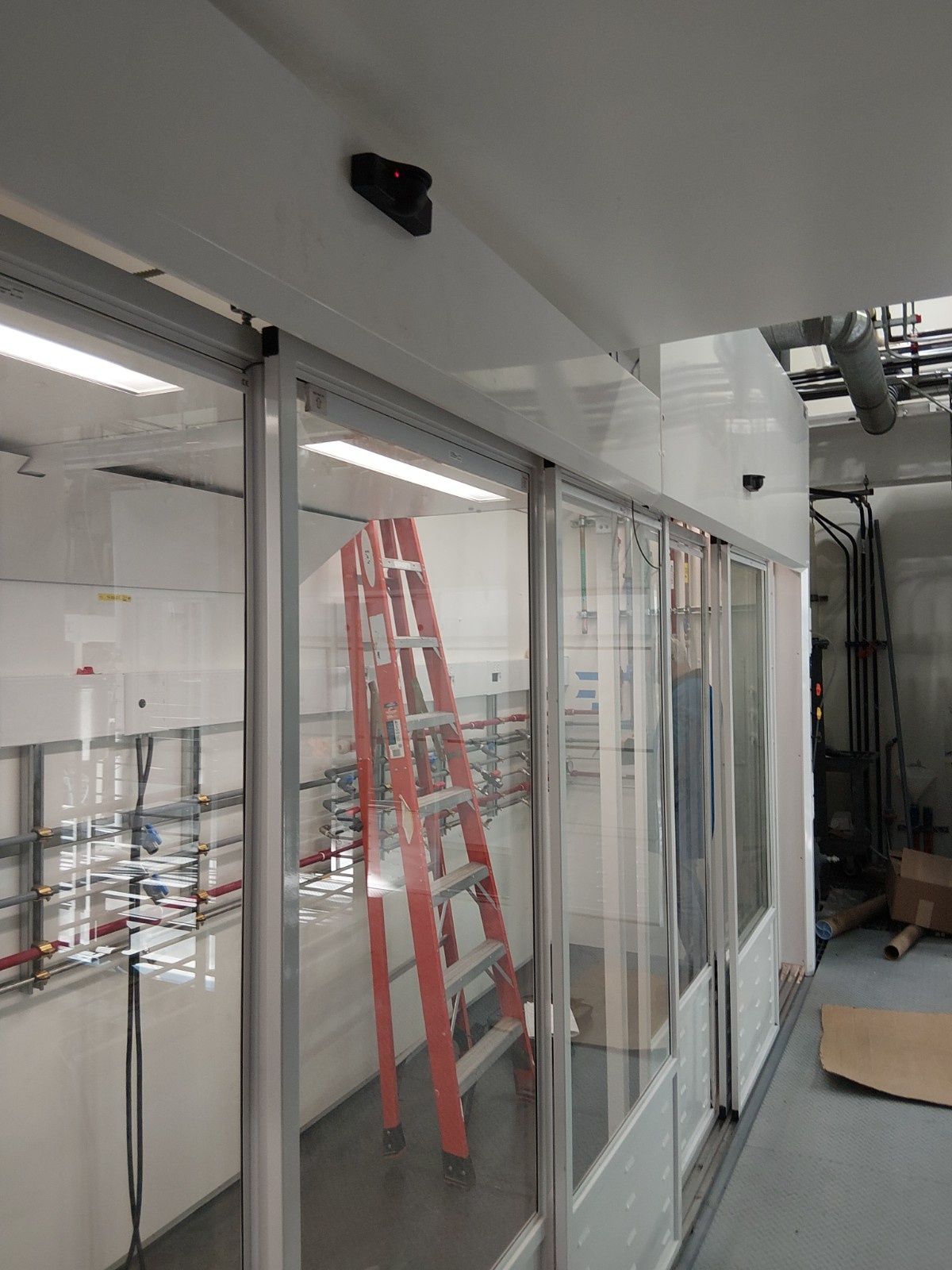 An orange A-frame ladder stands inside a white, glass-walled room enclosure during construction or maintenance.