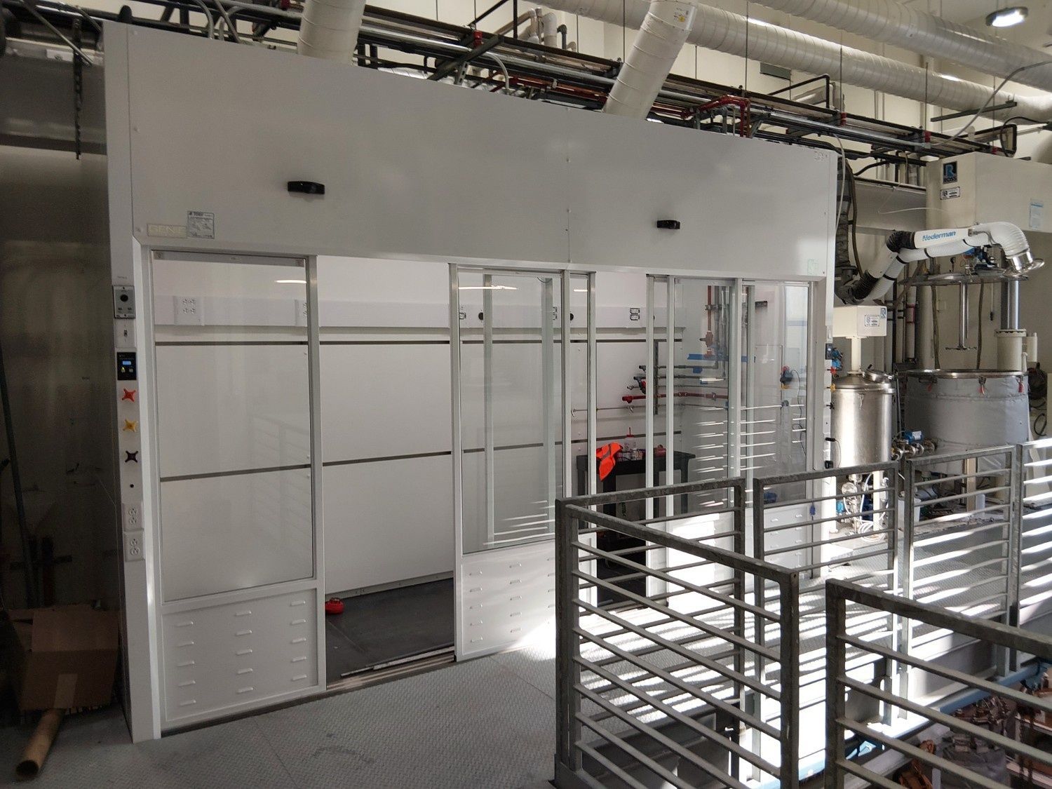 A white industrial laboratory enclosure with glass sliding doors sits on a platform next to stainless steel equipment.