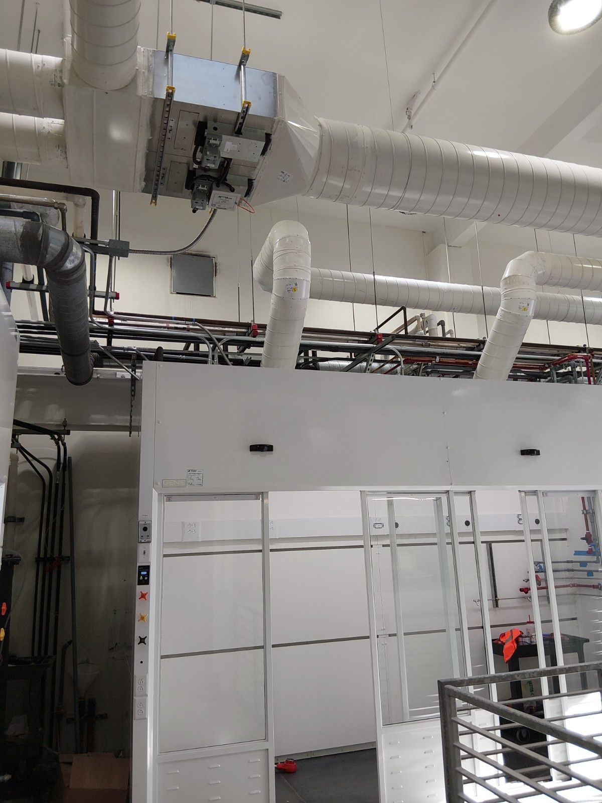 A lab area with white equipment racks, hanging ventilation ducts, and exposed pipes near a ceiling.