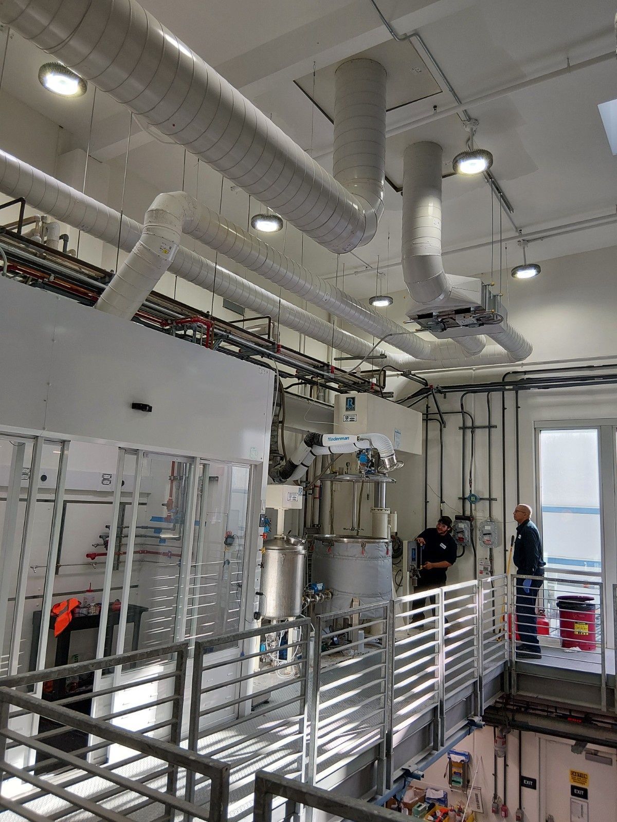 Two people work on industrial machinery in a high-ceilinged room with overhead ductwork and a metal walkway.