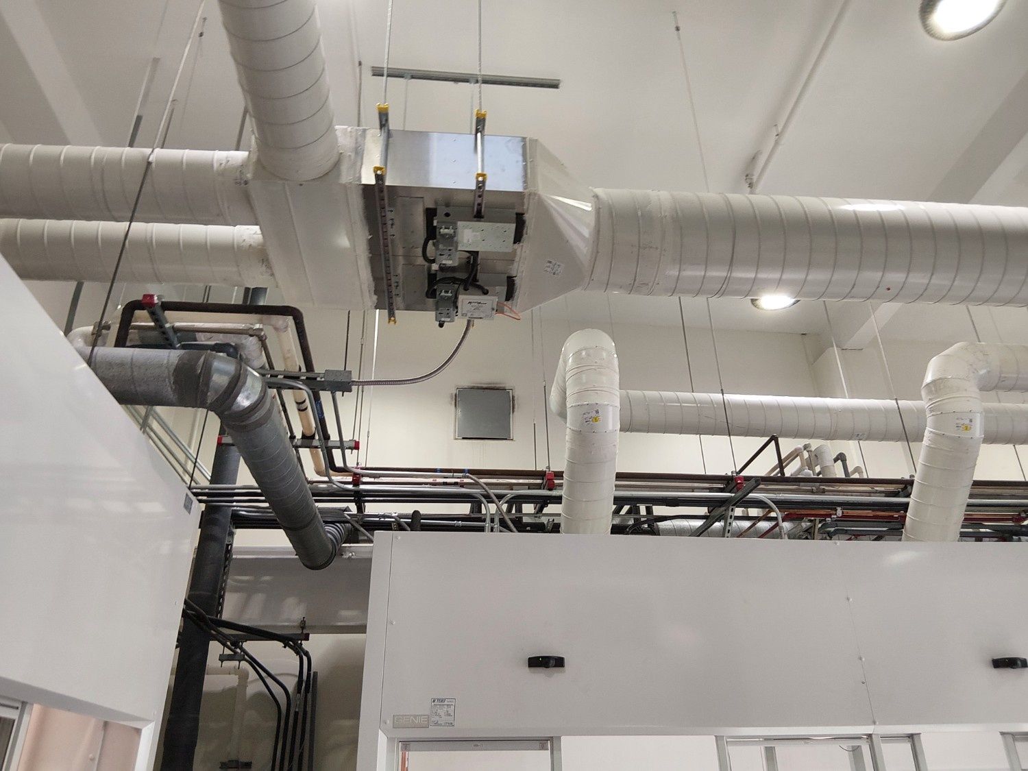 Industrial ceiling space showing HVAC ductwork, insulated pipes, and metal conduits suspended from above.