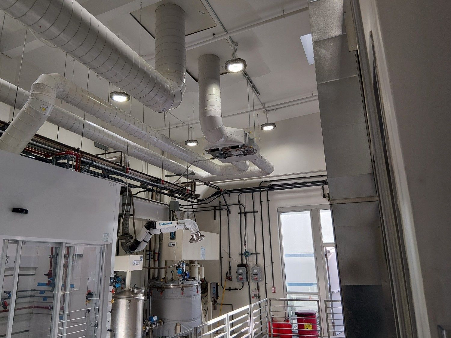 A technical lab space with exposed silver ductwork, overhead lighting, stainless steel equipment, and a robotic arm.