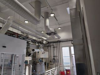 A laboratory interior featuring industrial machinery, silver stainless steel equipment, and overhead ventilation ducts.