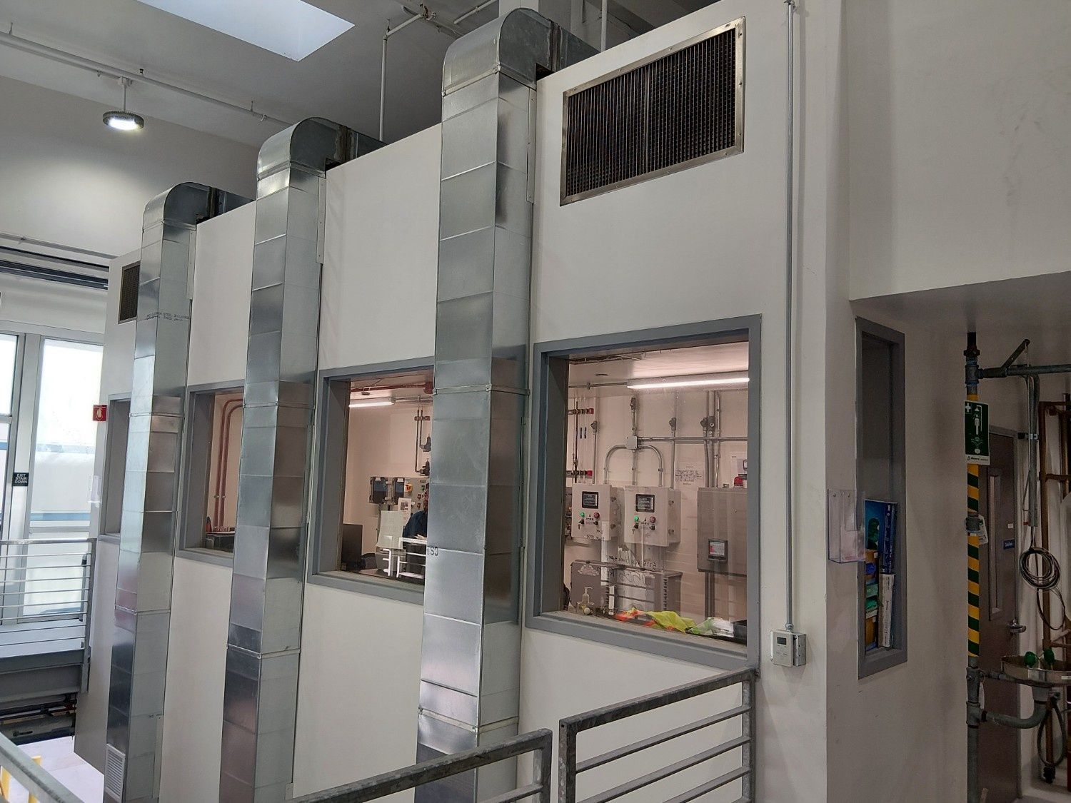 A white industrial wall with large windows, metal ventilation ducts, and a vent, inside a utility or laboratory space.