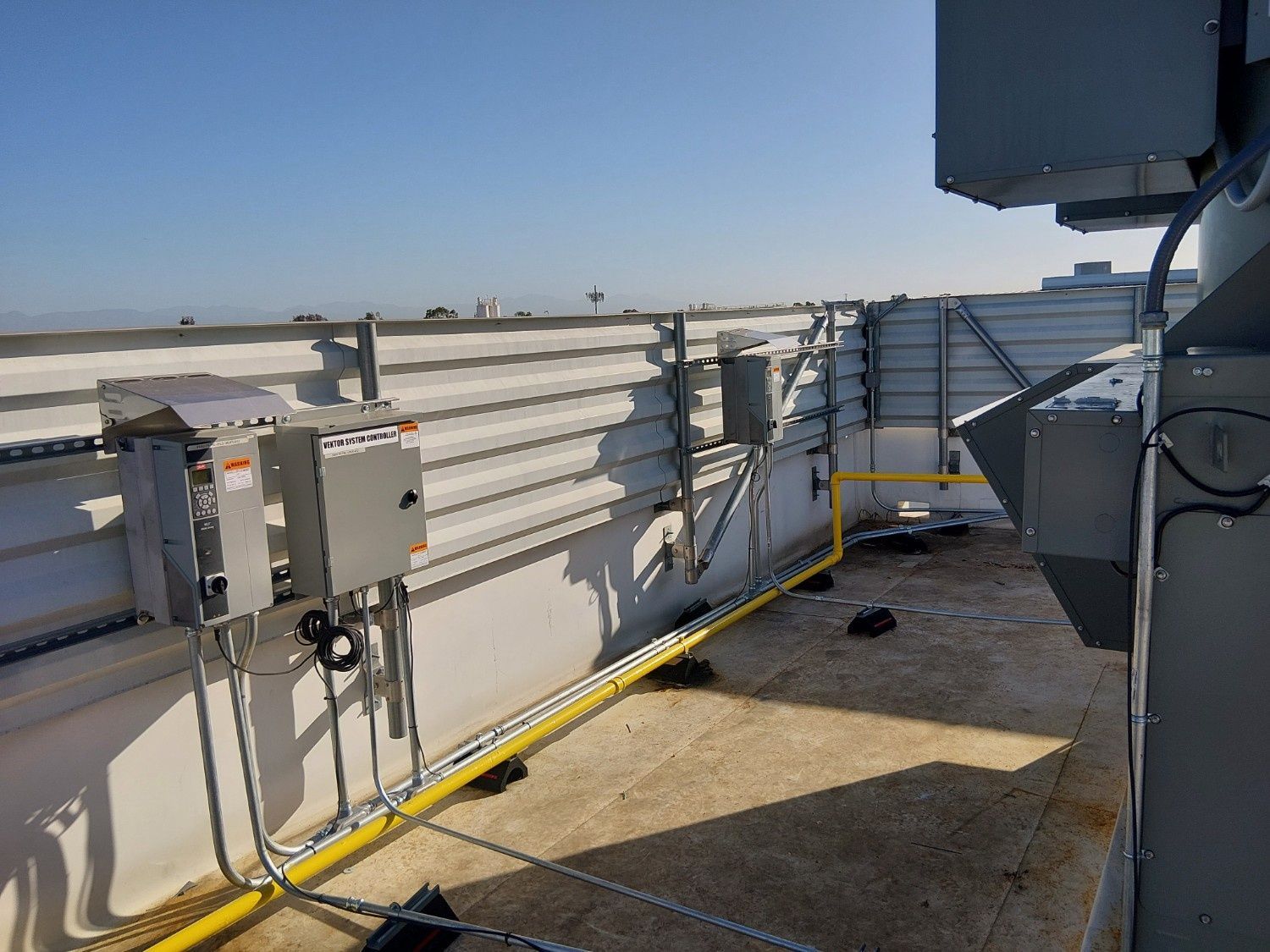 Electrical equipment and yellow gas piping installed along a metal roof railing against a clear blue sky.