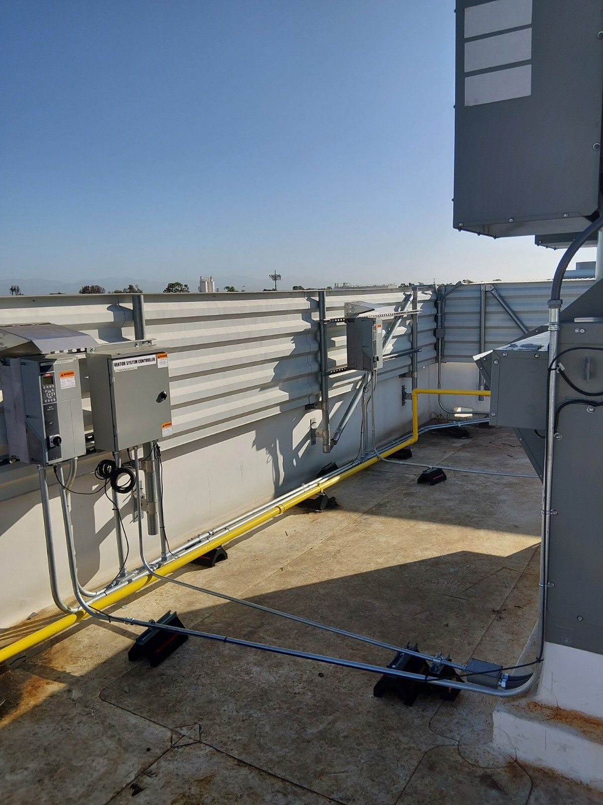 Electrical conduit and yellow gas pipes run along a flat rooftop near equipment boxes against a corrugated wall.