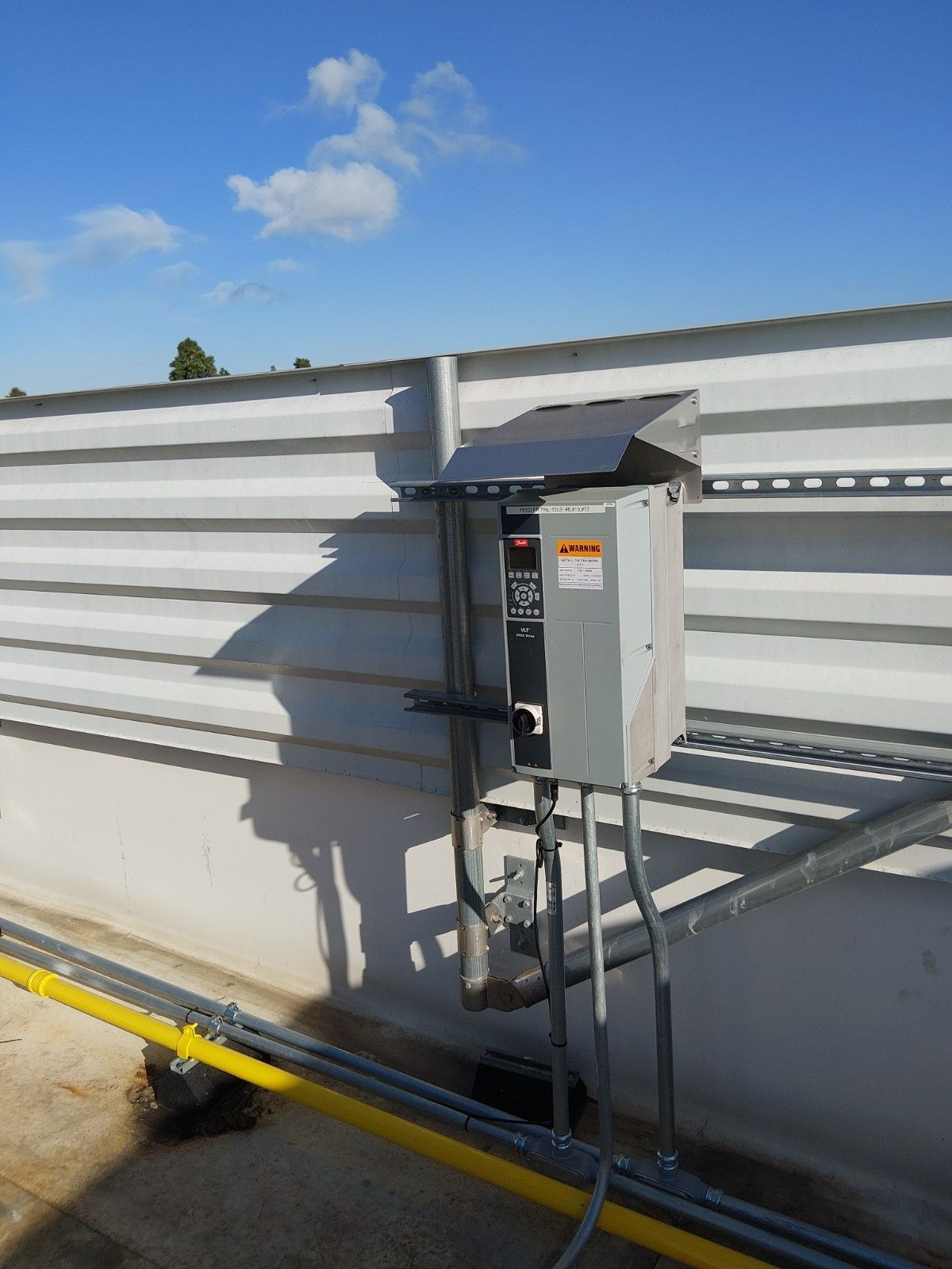 A gray electrical disconnect box mounted to a metal post against a corrugated wall under a blue sky.