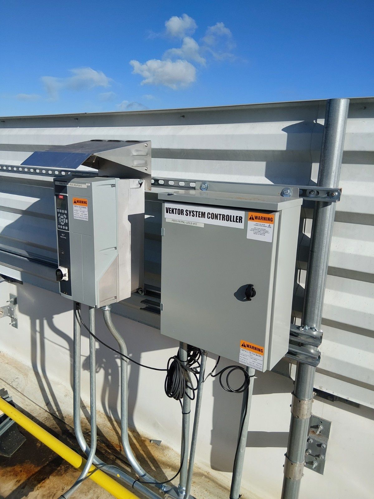 Two grey electrical enclosures mounted on a metal wall outdoors under a blue sky.