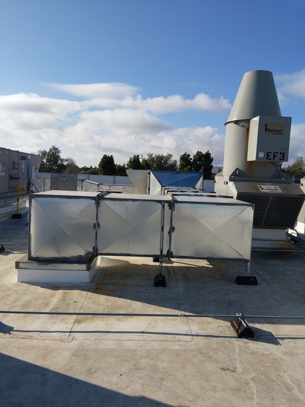 A rooftop ventilation unit with a tall, grey exhaust fan and connected rectangular metal ductwork under a clear sky.