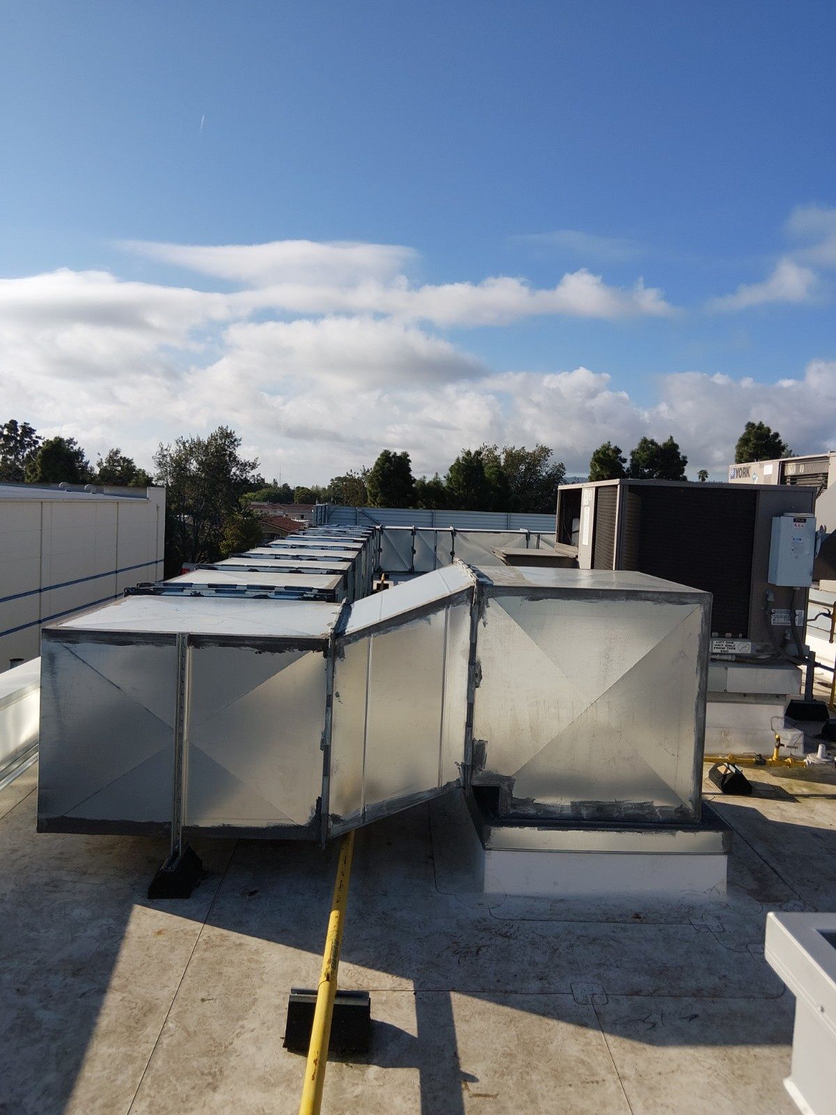 A row of square metal HVAC ducts on a flat roof under a blue, cloudy sky.