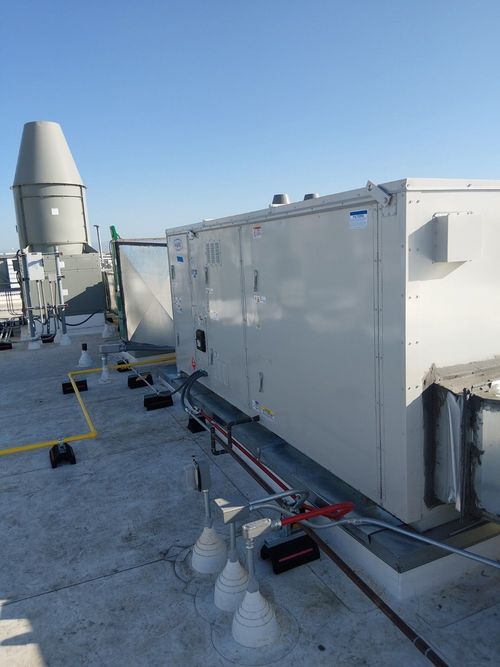 A rooftop HVAC unit with light-colored panels, piping, and a large metal exhaust vent under a clear blue sky.