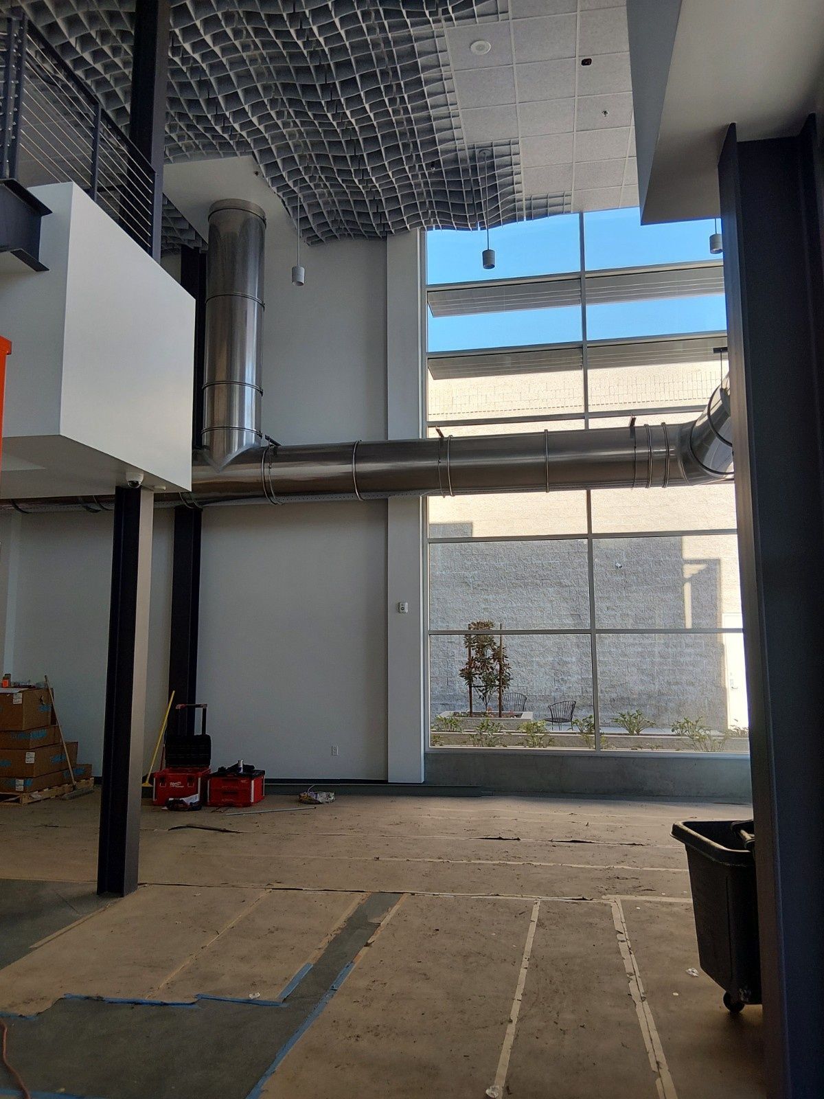 An indoor construction site features a high ceiling with metal ductwork, tall windows, and an unfinished concrete floor.