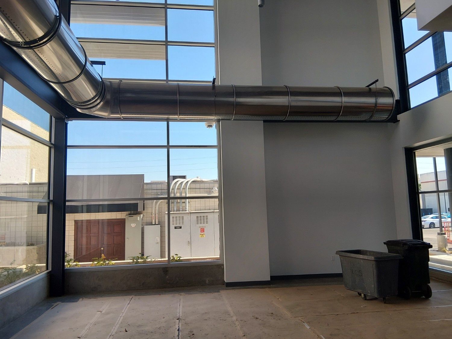 An interior view of a modern space with large windows, a metal duct across the ceiling, and two black bins on the floor.