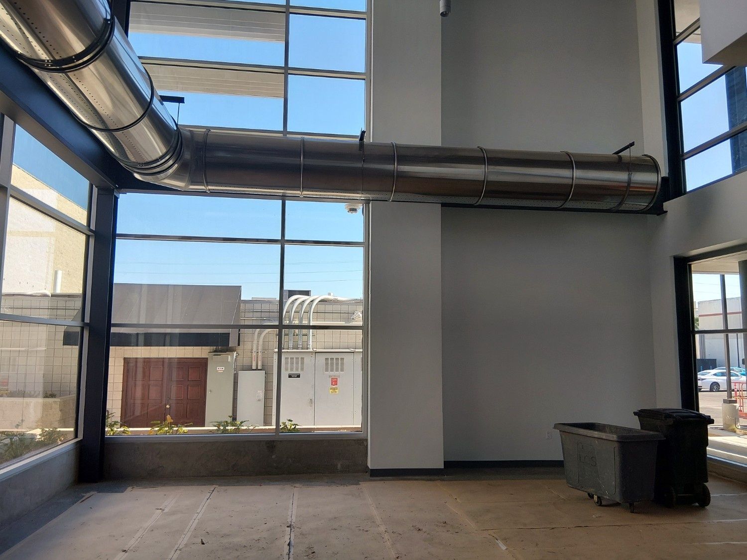 A modern, empty room featuring floor-to-ceiling windows, exposed industrial ductwork, and two large waste bins.