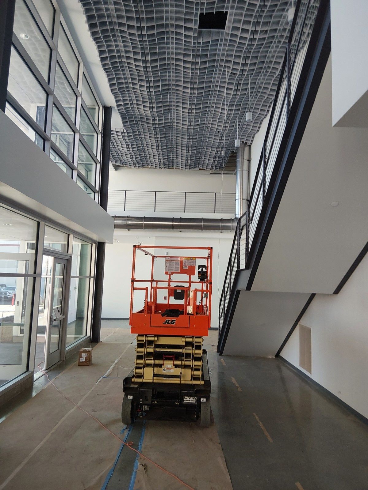 An orange scissor lift sits in the center of an indoor lobby with large windows and a staircase under a ceiling grid.