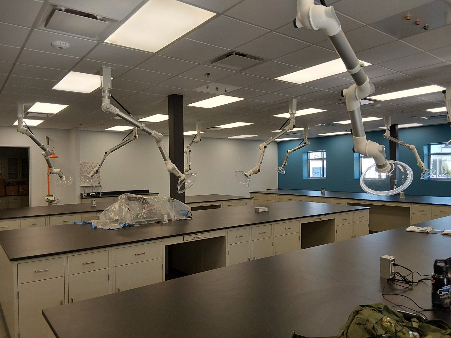 A science laboratory with multiple workstations, light-colored cabinets, dark countertops, and flexible ceiling extractors.
