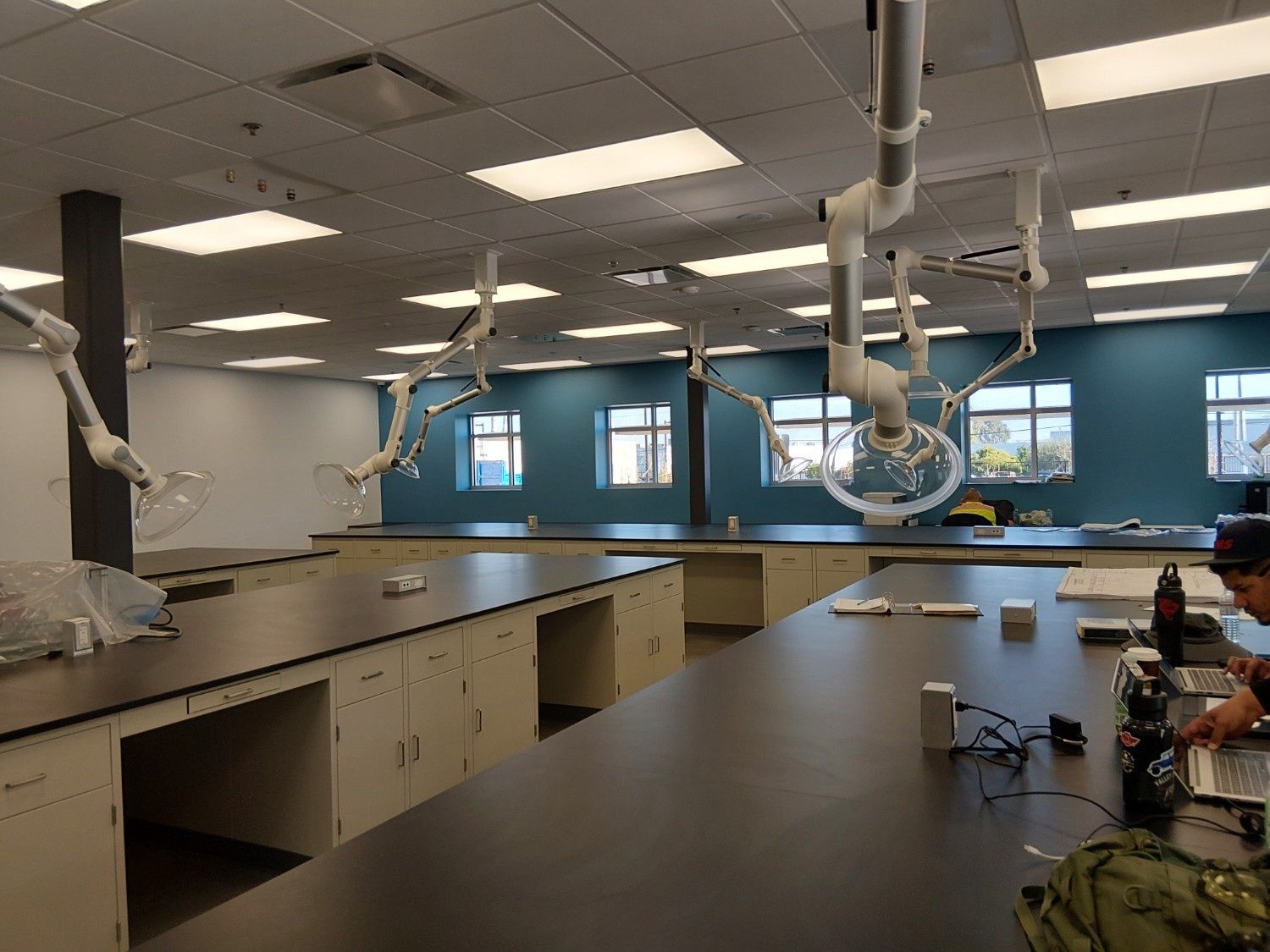 A lab room with rows of long tables, white fume extractor arms, and a person working on a laptop at a table.