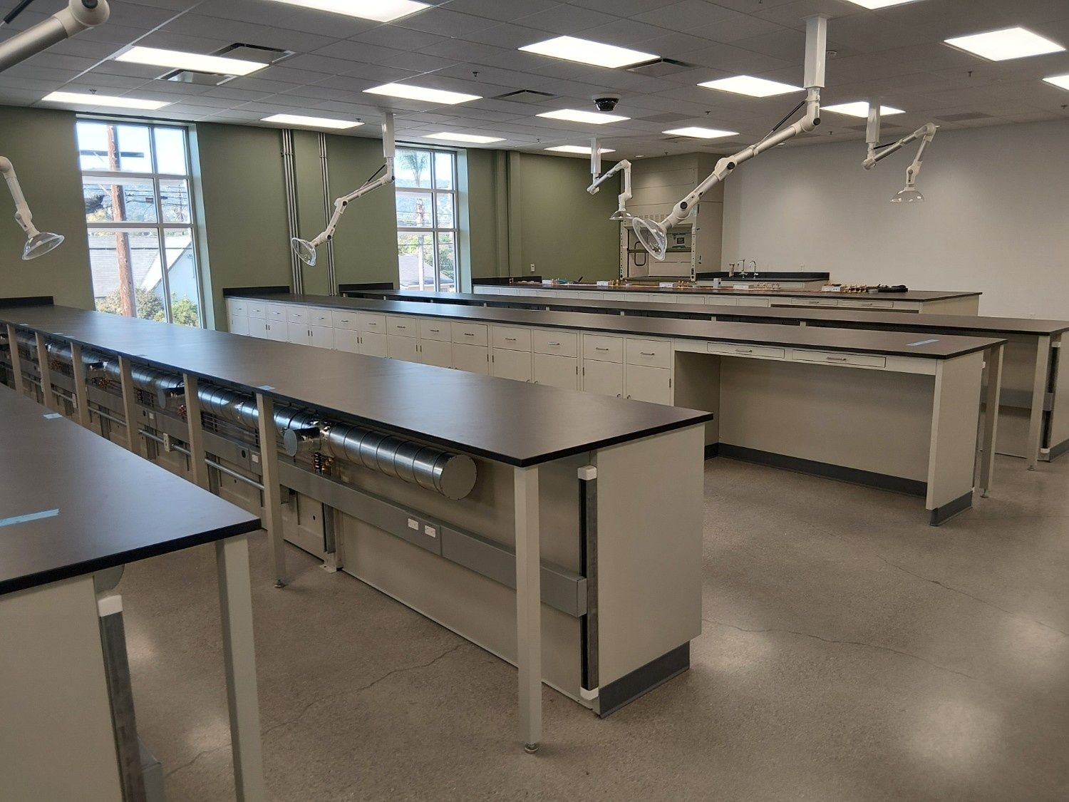 Empty laboratory classroom featuring rows of workstations with cabinets, black tabletops, and overhead ventilation arms.