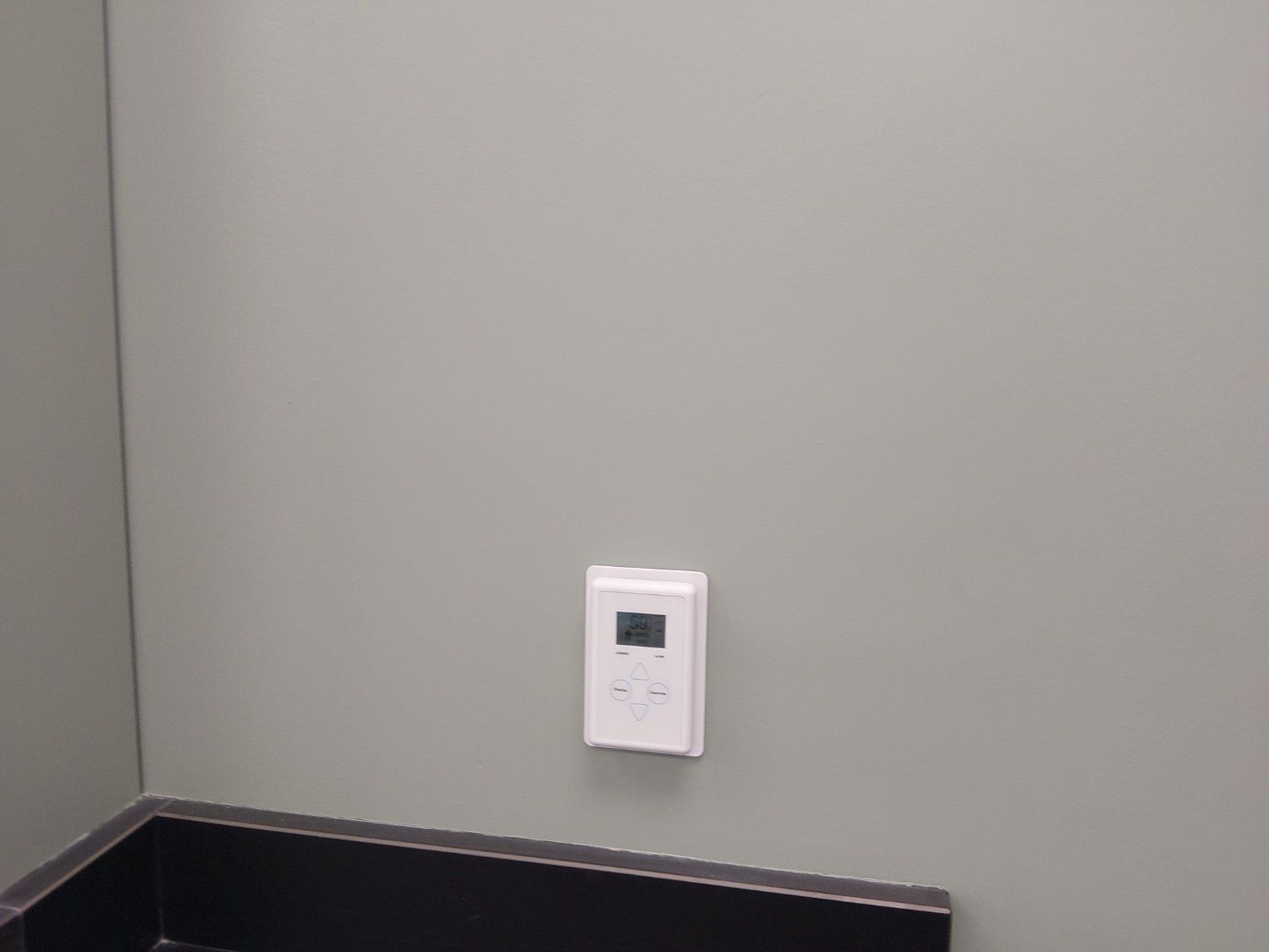 A white digital wall-mounted thermostat control panel centered on a light gray wall above a black surface.