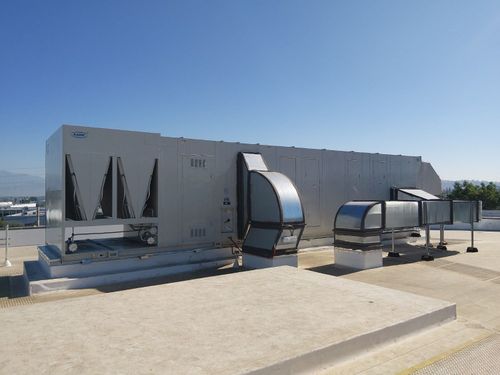 A large gray rooftop HVAC unit with silver ductwork stands under a clear blue sky on a flat gravel roof.