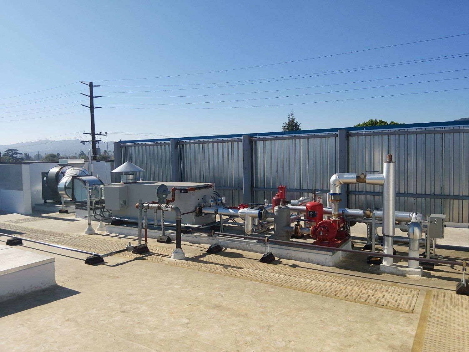 Industrial HVAC equipment with silver metal piping and red pumps on a flat gravel rooftop against a blue sky.