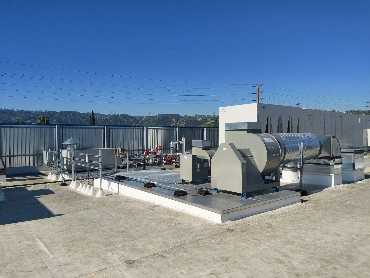 Industrial equipment on a flat rooftop, featuring a large gray ventilation blower and metal pipes against a blue sky.