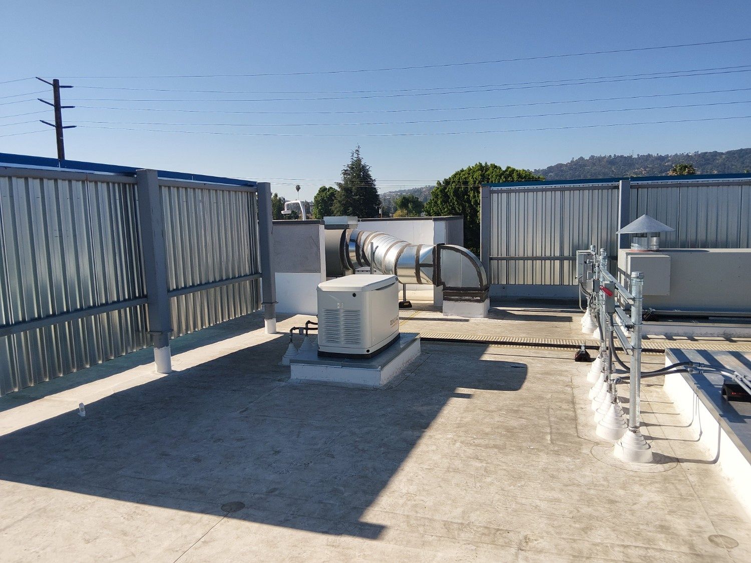 A rooftop scene showing a generator, metal ductwork, and safety railings against a clear blue sky.