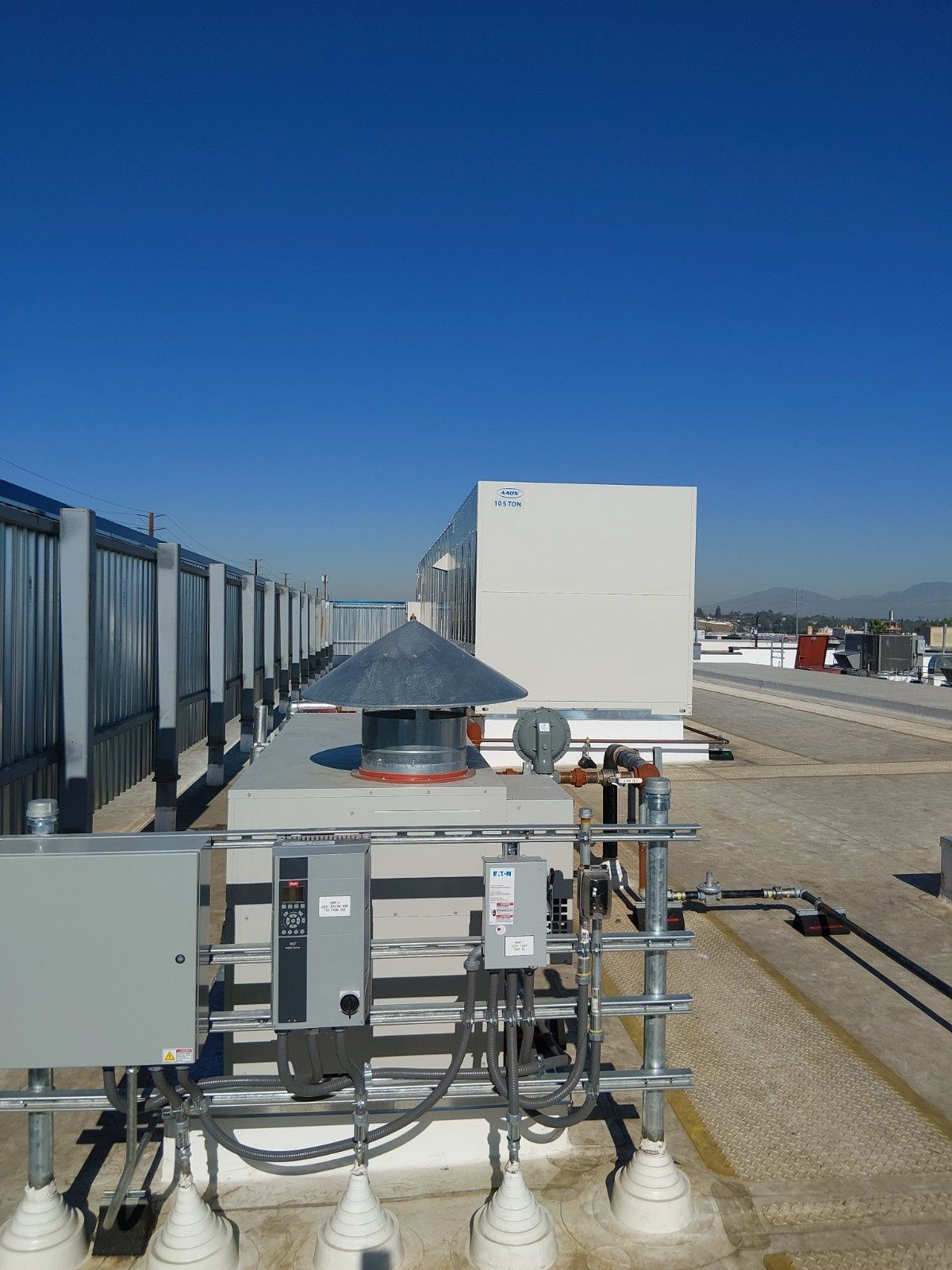 Industrial rooftop equipment includes a conical vent, electrical control boxes, and conduit against a clear blue sky.