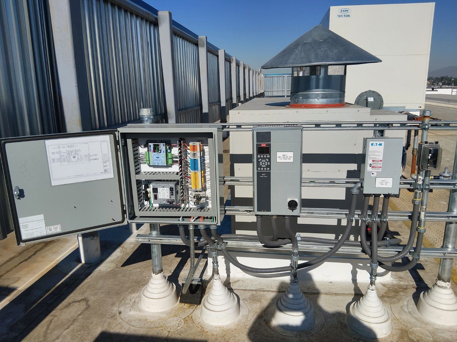 An open electrical control panel and variable frequency drive mounted on a rooftop railing near an exhaust vent.