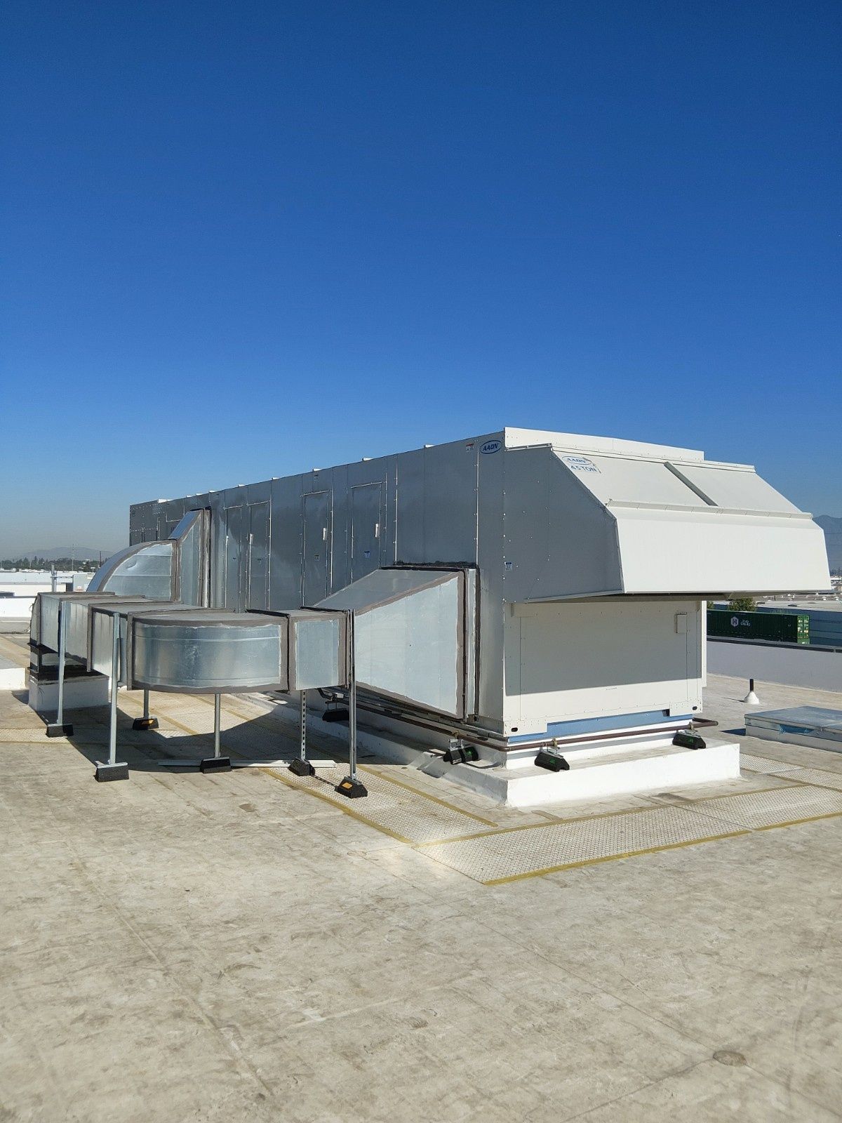 A large industrial HVAC unit with silver metal ductwork sits on a flat rooftop against a clear blue sky.