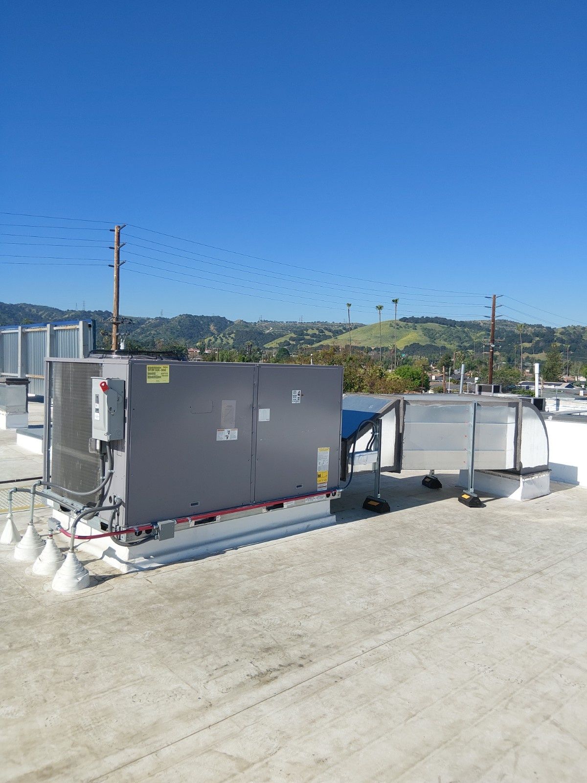A grey commercial HVAC unit sits on a flat, gravel-covered roof against a clear blue sky and distant hills.