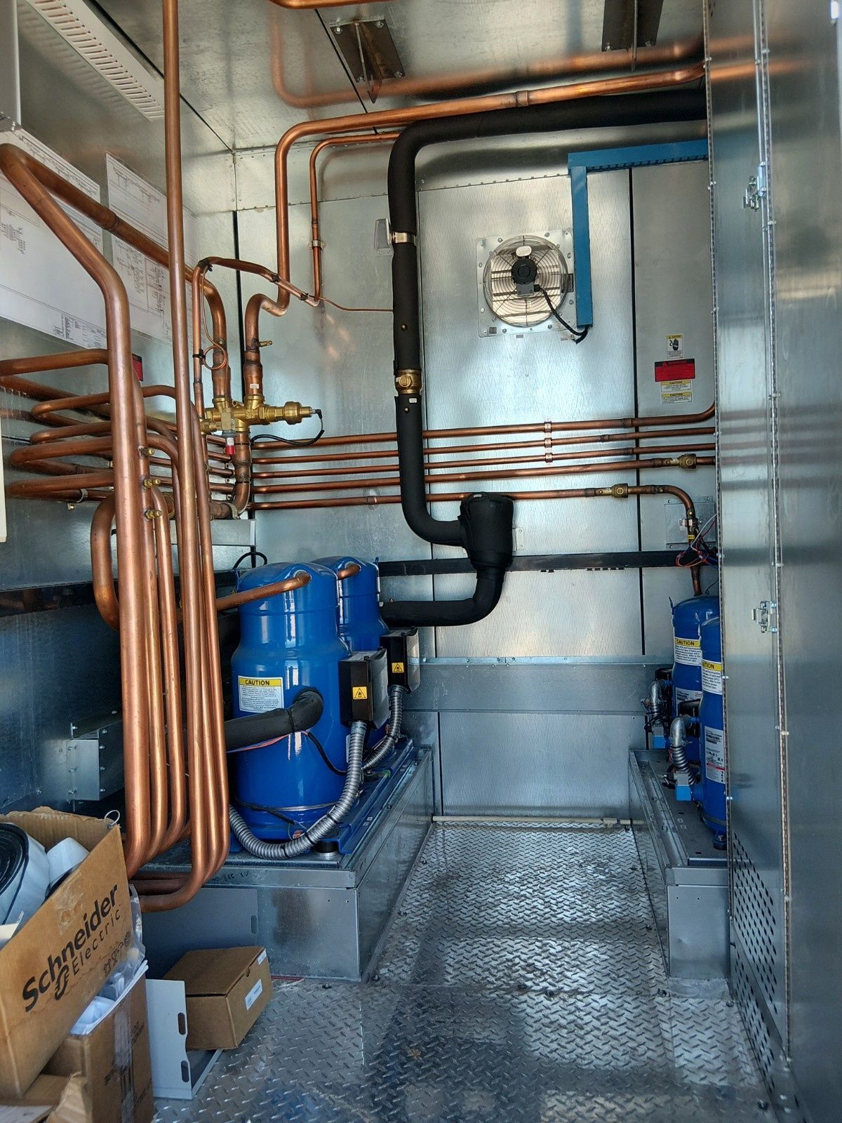 Interior view of a mechanical room featuring blue industrial compressors, copper piping, and black insulation on walls.