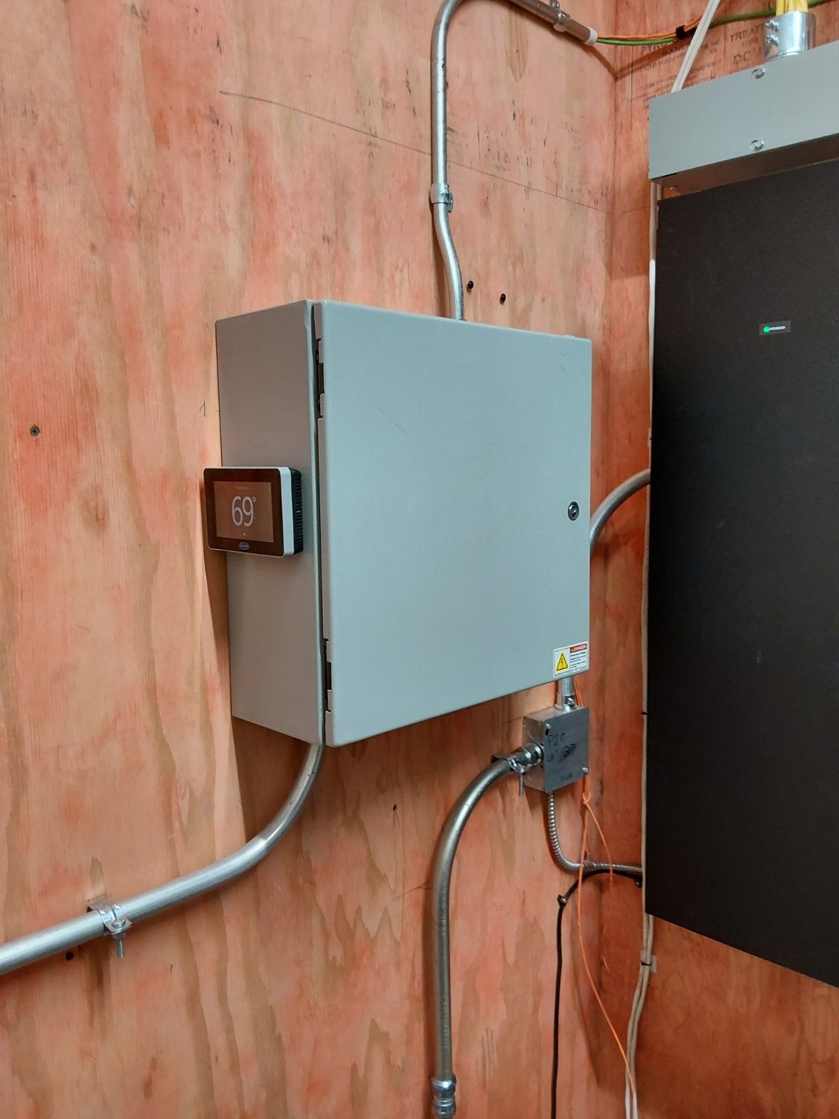 A grey electrical control box mounted on a plywood wall next to a large black panel with conduits running to it.