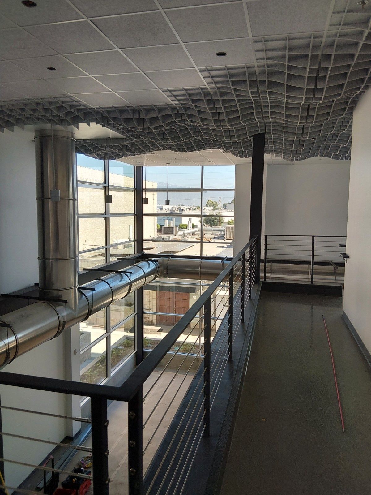 A hallway with a metal railing overlooks a sunlit atrium featuring a unique, textured, cloud-like ceiling installation.