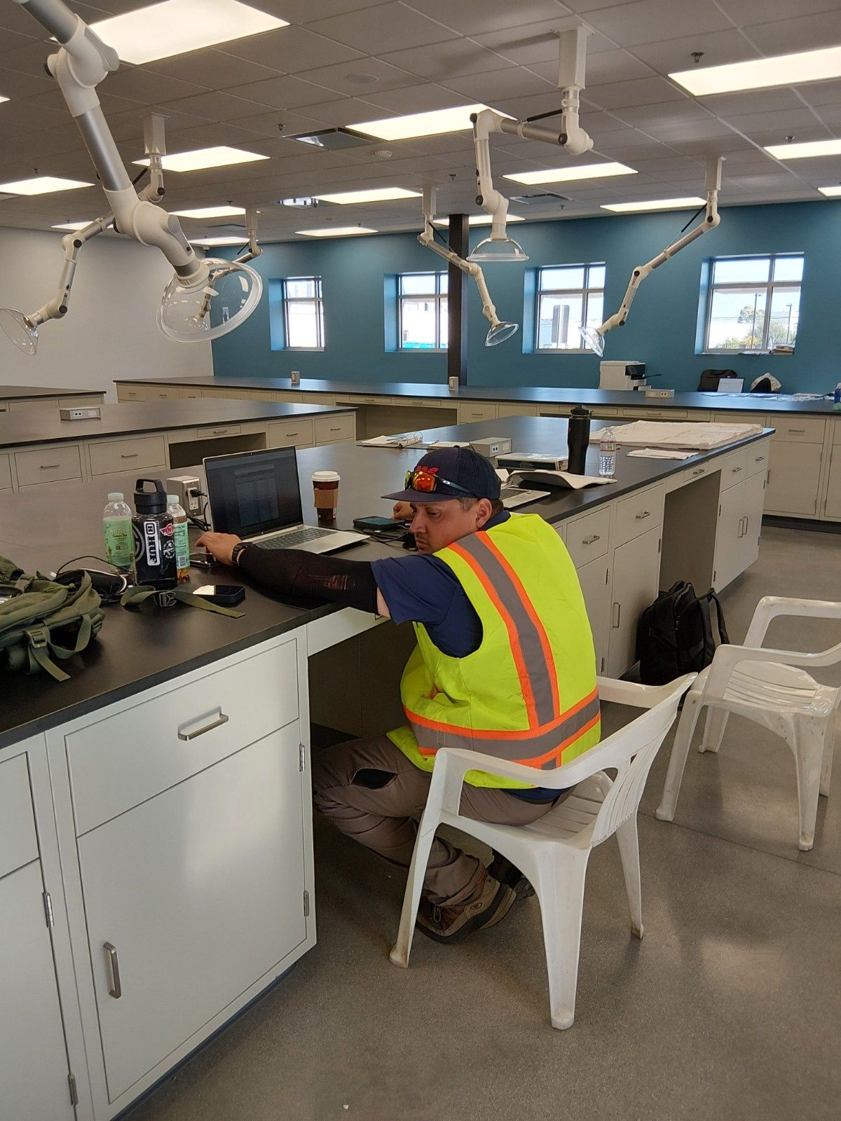 A person wearing a high-visibility vest sits at a workbench in a lab, working on a laptop.