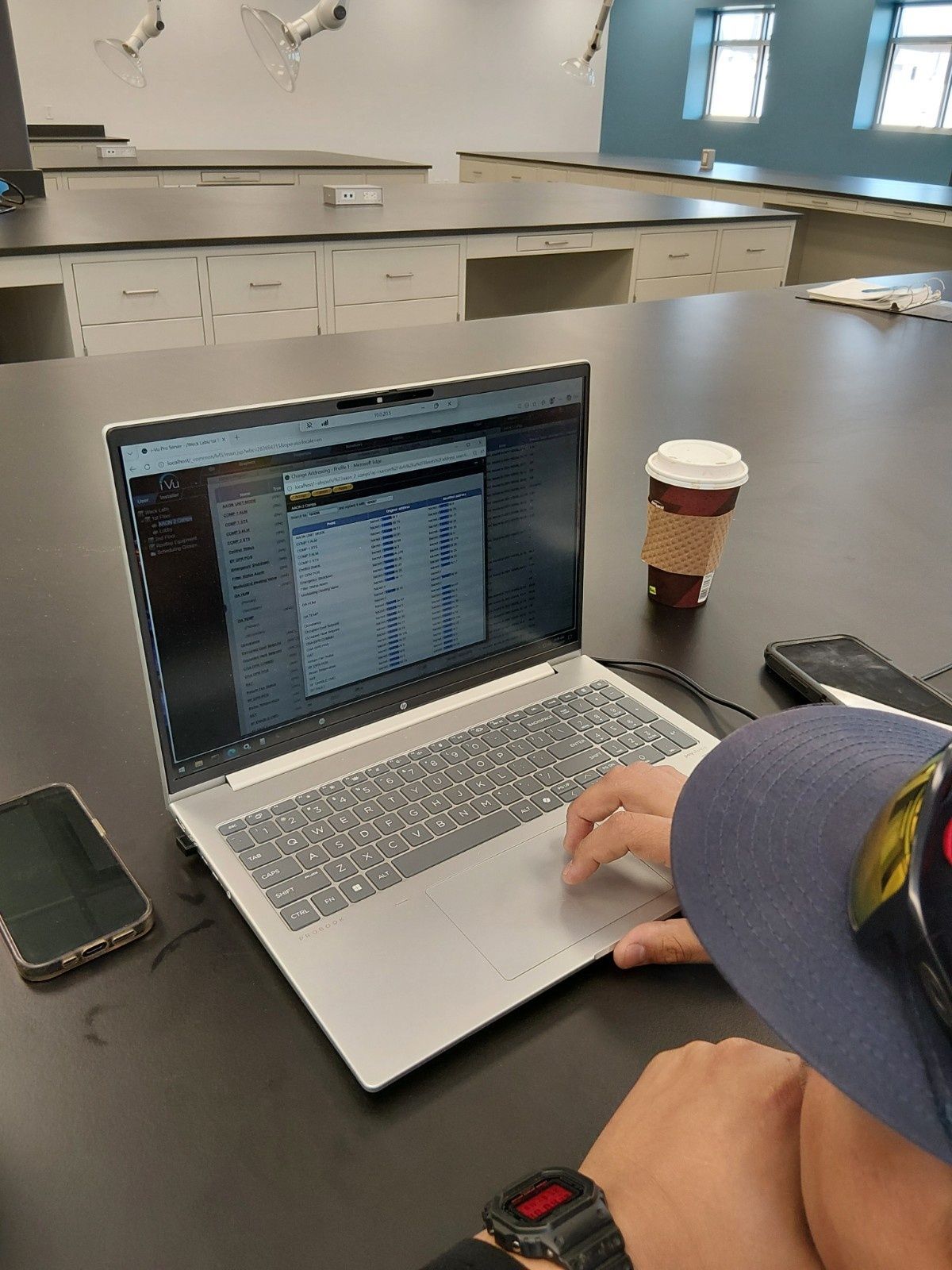 A person uses a laptop displaying data tables at a large desk in a bright, modern classroom.