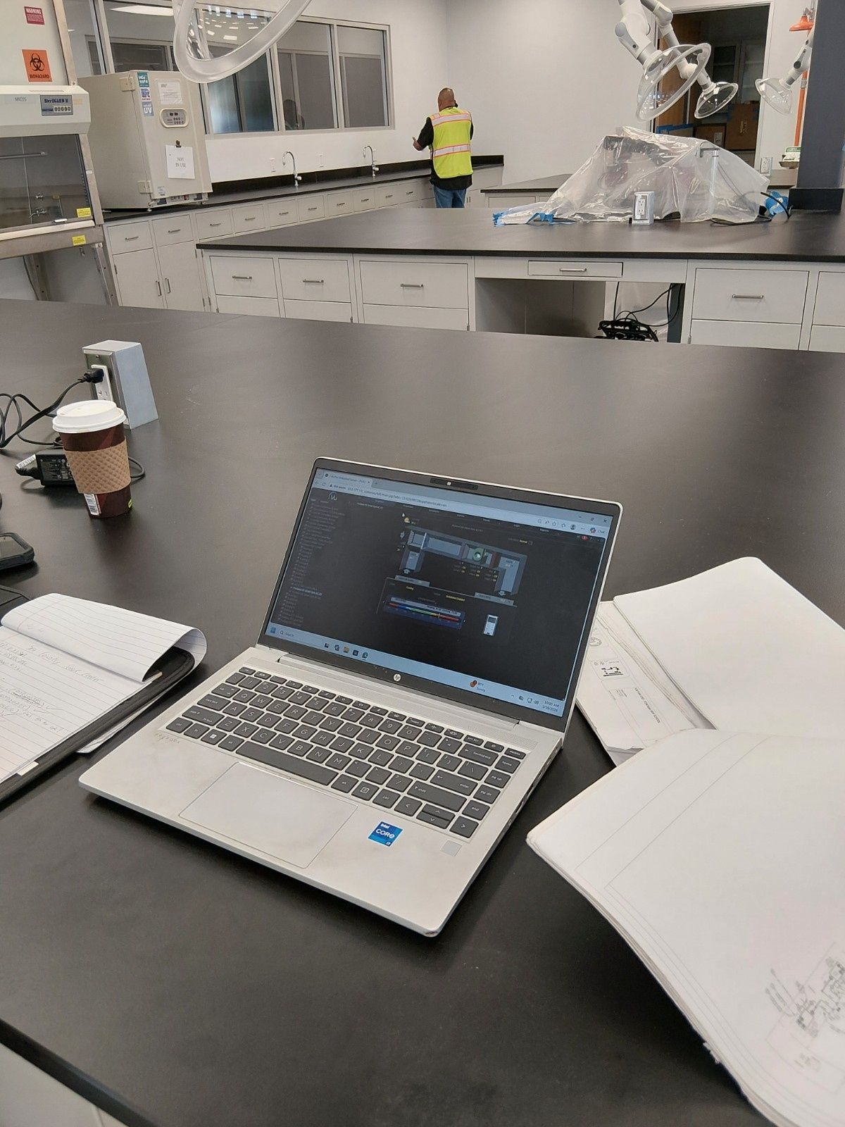 A laptop with a dark screen, a cup of coffee, and notebooks sit on a laboratory counter. A person works in the background.