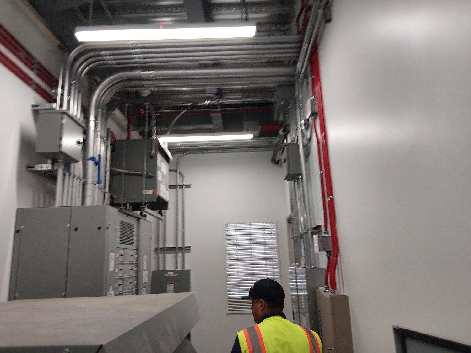 A worker in a high-visibility vest stands in a utility room with metallic conduit pipes and electrical control panels.