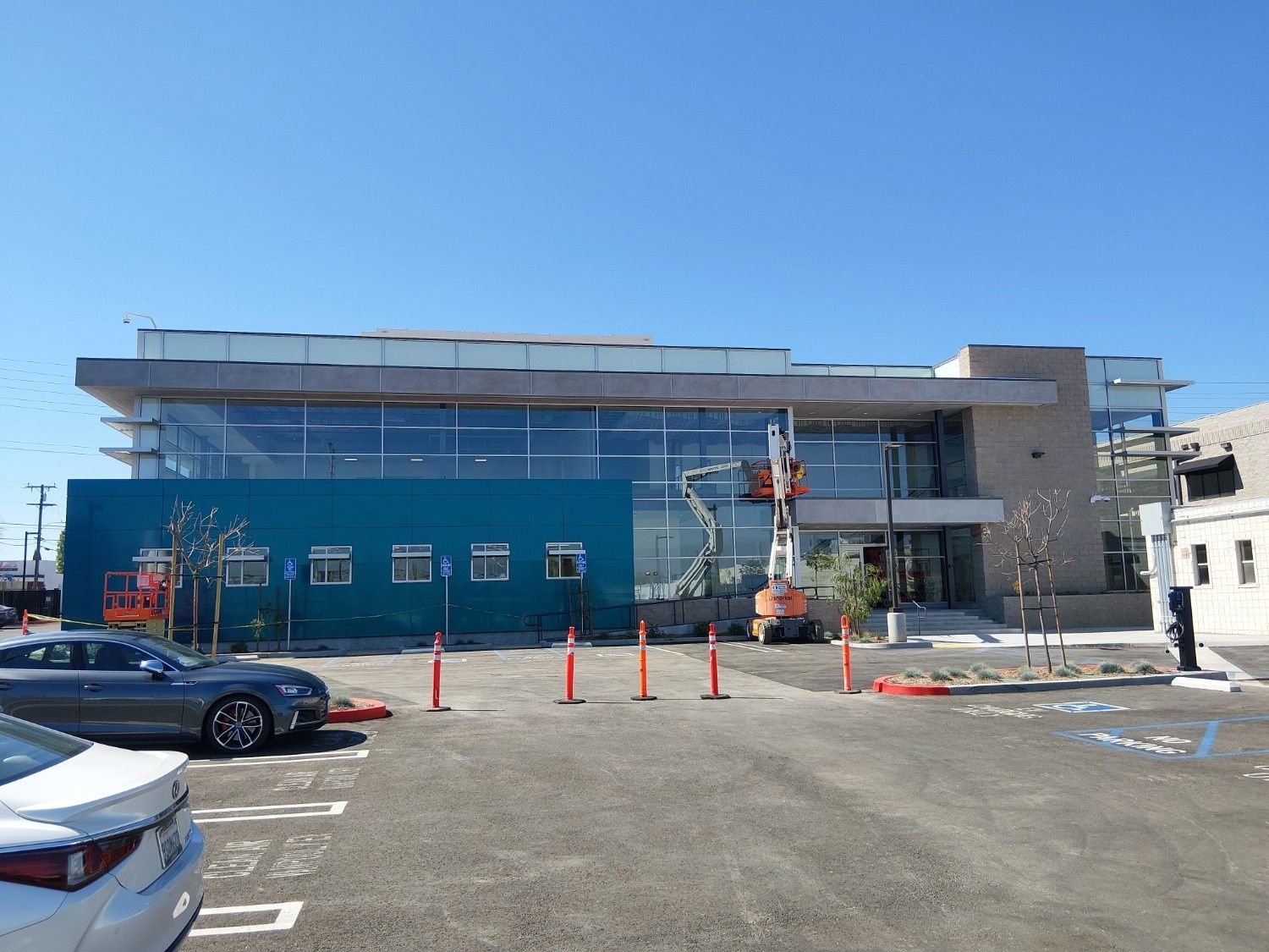 Modern office building with a teal facade, glass windows, and a construction lift in a sunny parking lot.