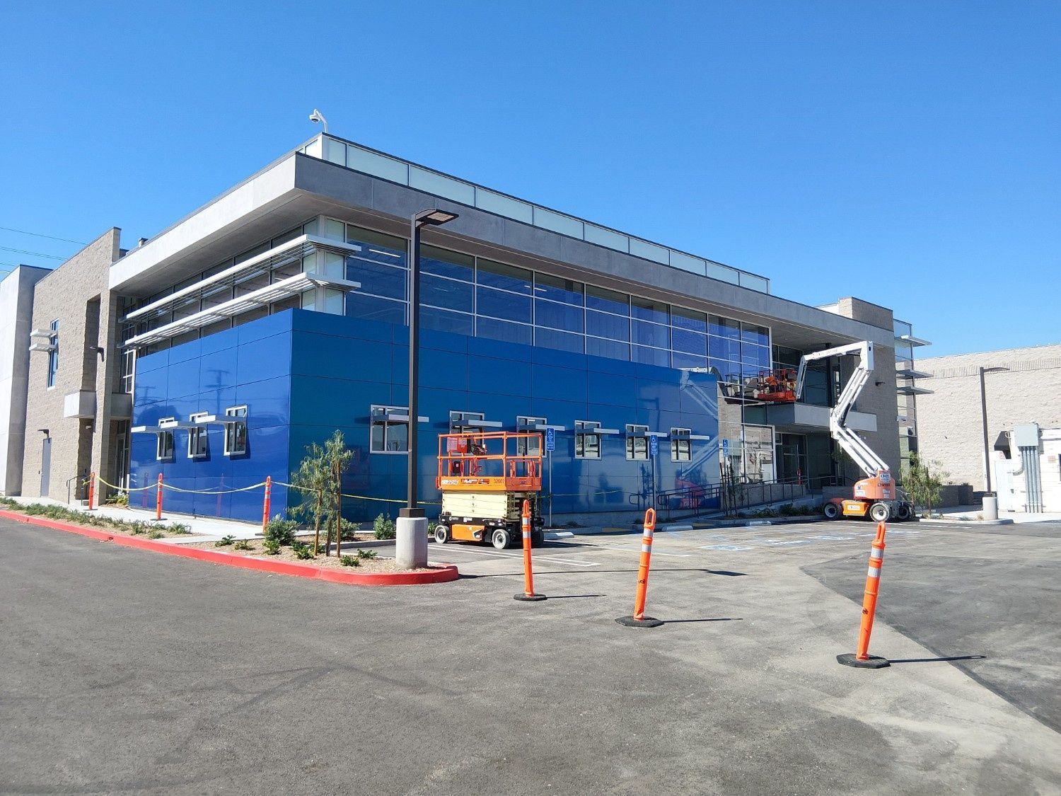 A modern building under construction features a large blue glass facade, an orange scissor lift, and a boom lift.