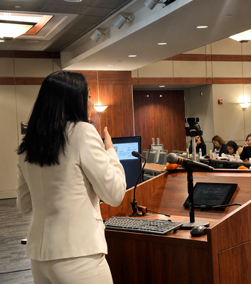 Speaker at a podium in a conference room, addressing an audience with a laptop and microphone nearby.