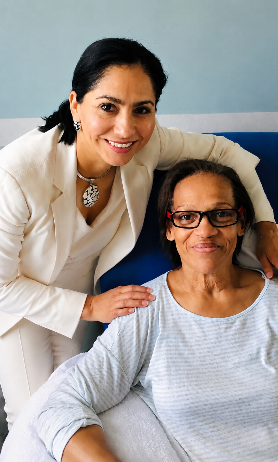 A person in a light blazer stands behind another person sitting down, placing a hand on their shoulder. Both are smiling.