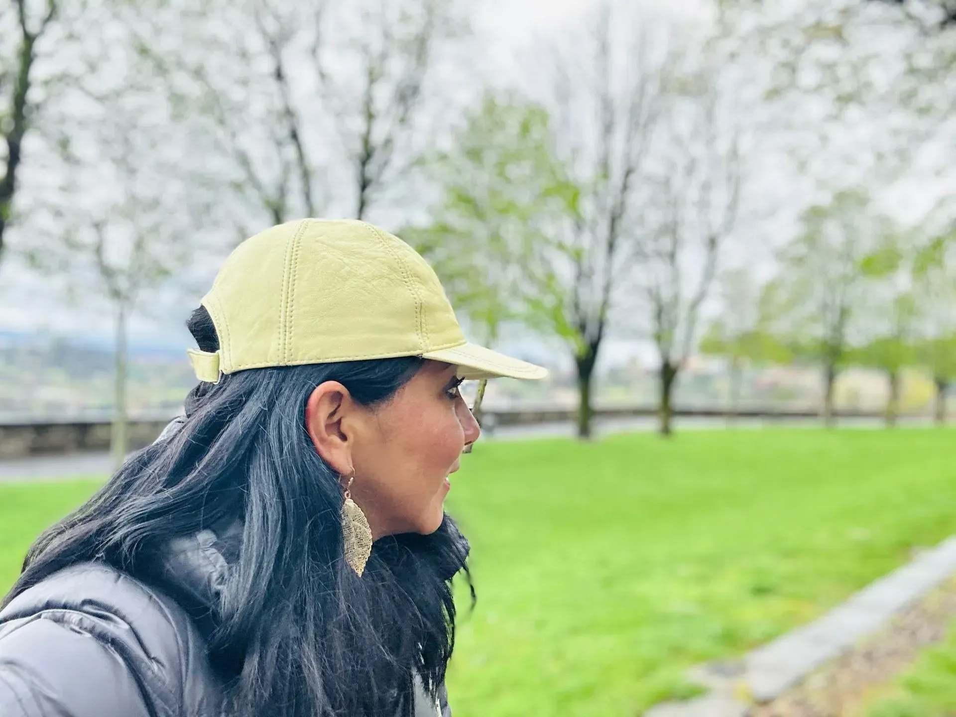 Woman with dark hair, wearing a yellow cap and earrings, looking towards green space and trees.