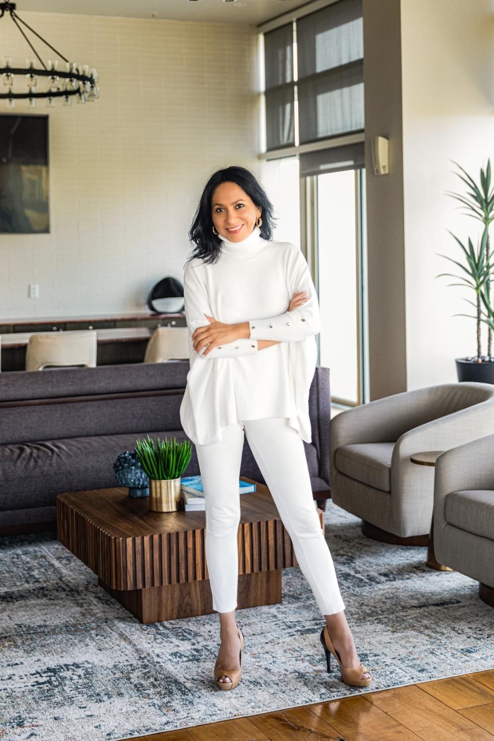 Woman in white outfit stands in modern living room with arms crossed.
