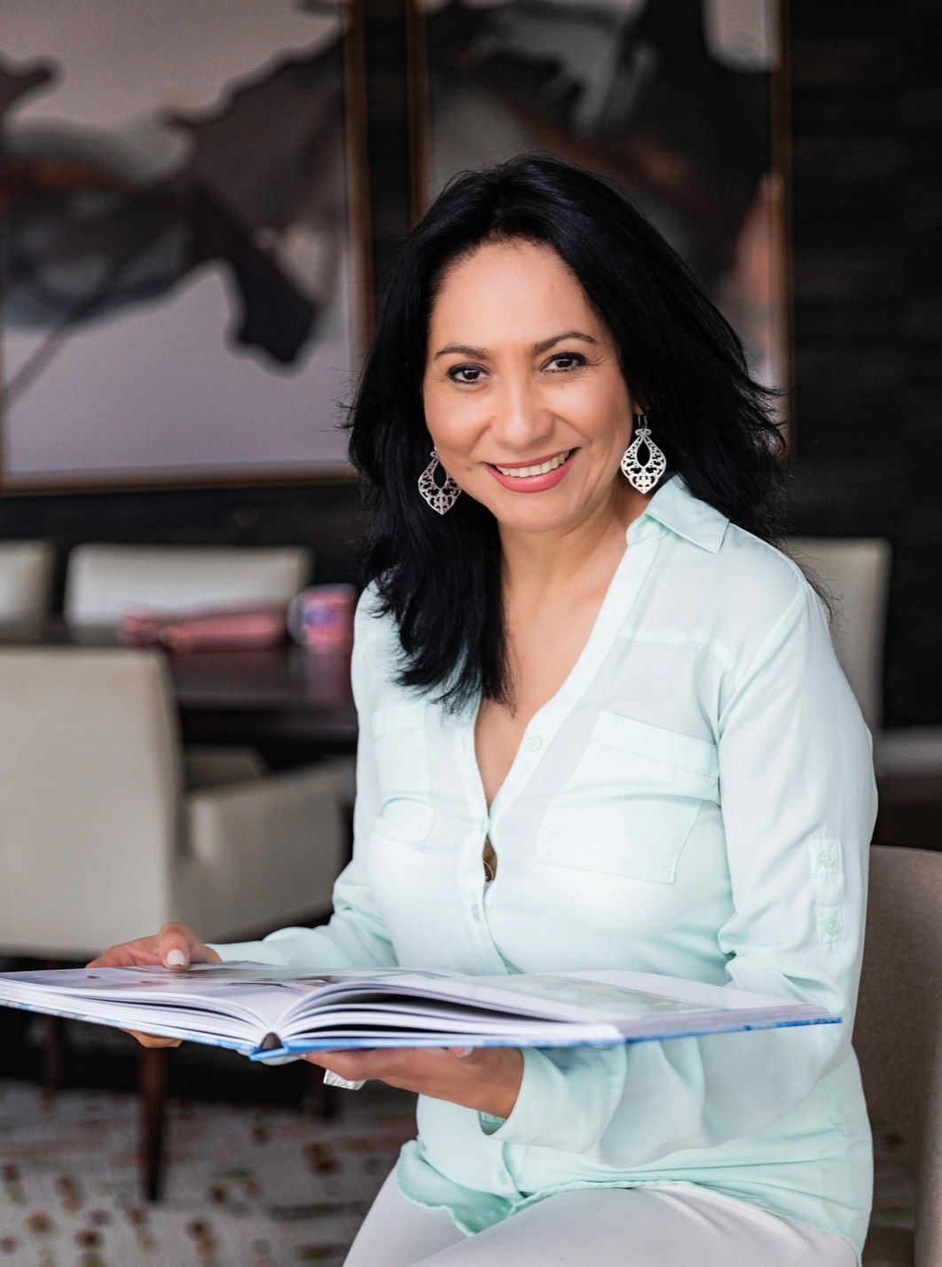 Woman smiling, holding a book, seated in a well-lit room. She is wearing a light blue shirt.
