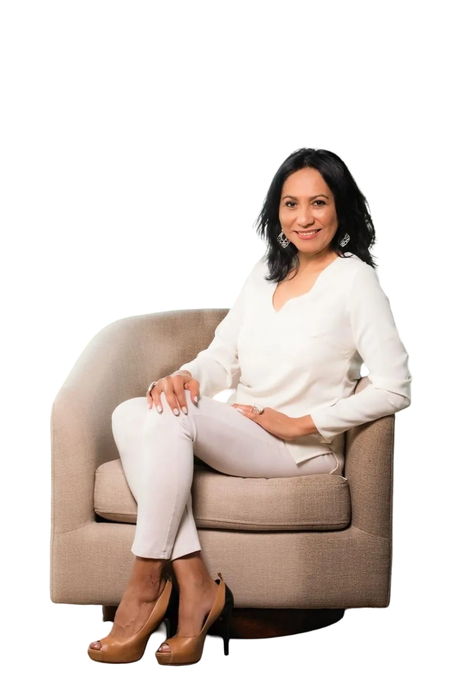 A smiling individual sitting in a neutral-colored, rounded chair, wearing a white long-sleeved top, white pants, and heels.