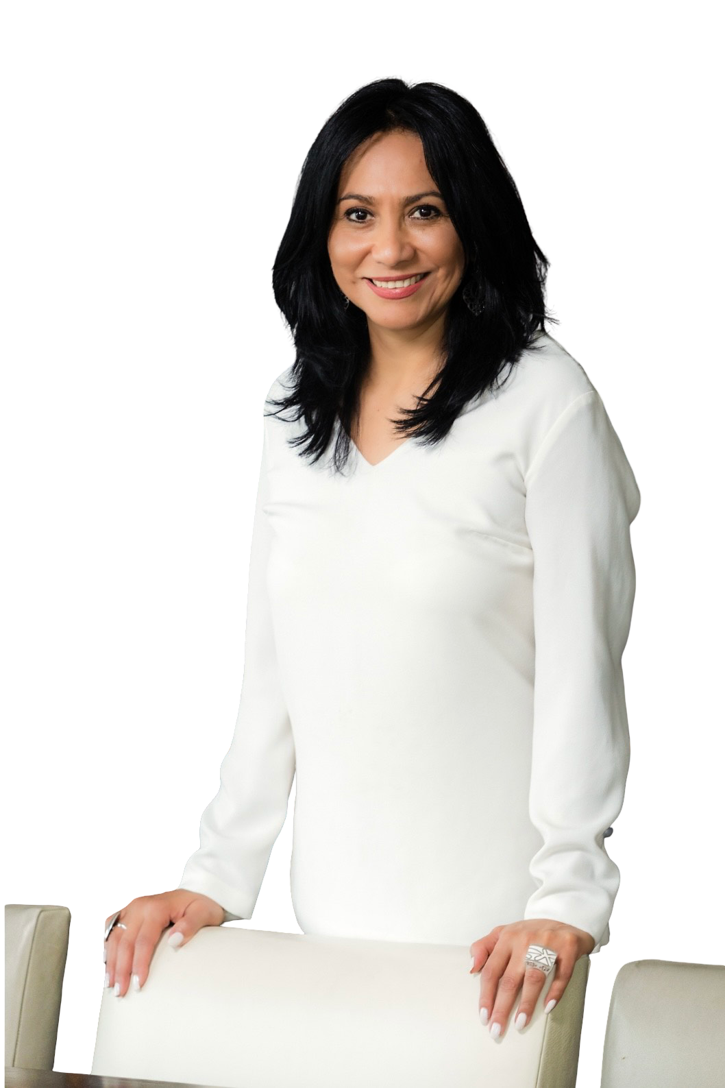A smiling person with dark hair wears a long-sleeved white top, standing with their hands resting on a light-colored chair.