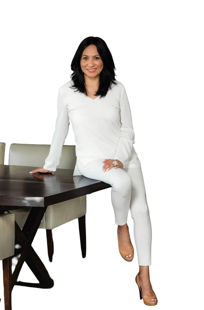 A person with dark hair wearing all-white clothing, sitting on the corner of a dark wooden dining table.