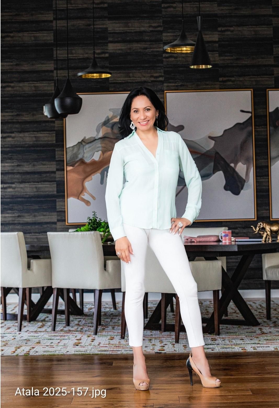 Woman in light blue shirt and white pants poses in a dining room with abstract art.