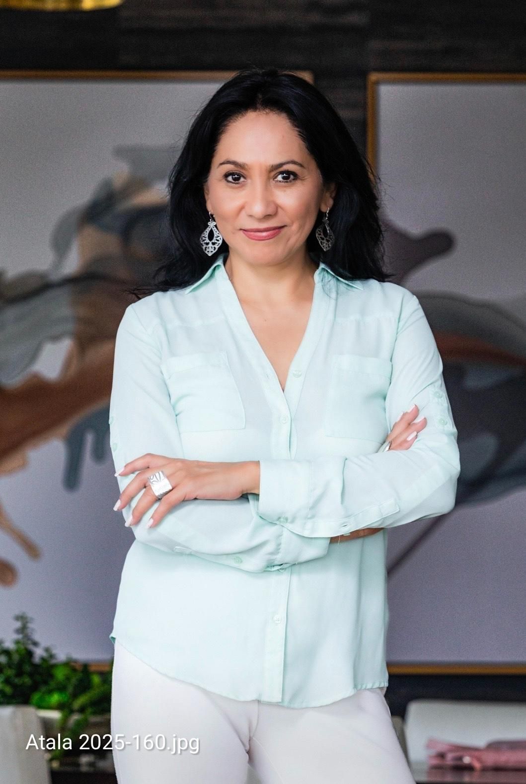 Woman in light blue shirt, arms crossed, smiling. White pants, silver earrings, and ring. Standing indoors near artwork.
